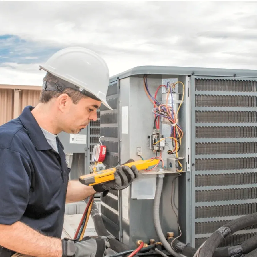 HVAC technician in a hard hat testing an outdoor air conditioner with a multimeter